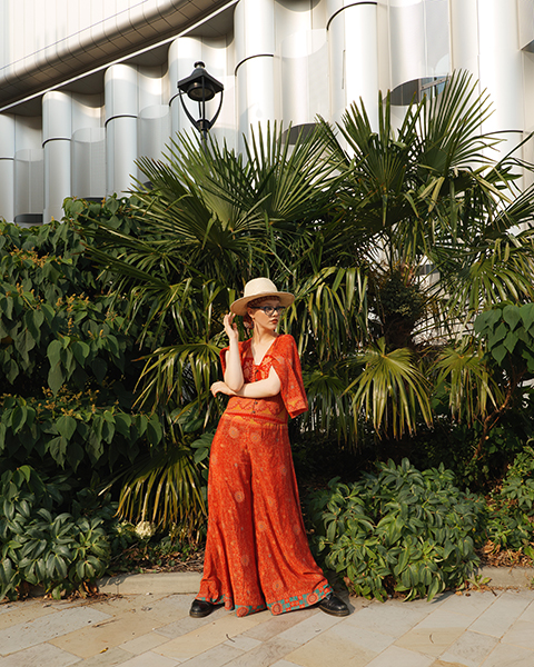 Female model posing stood in an orange jumpsuit infront of botanical hedge