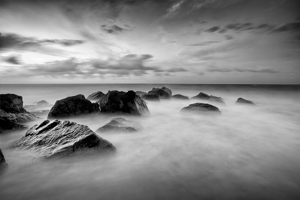 a black and white long exposure of the tide rolling out creating a finely blurred almost mist like effect around in stark contrast with the deep black jagged rocks that rise out of the tide pool