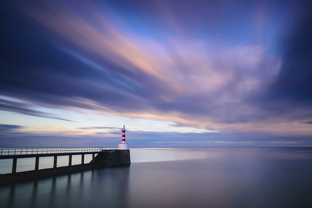 a striking image of a jetty reaching out in a perfectly straight line into the bay cutting the picture from the left to the centre of the frame, at the centre a red and white striped lighthouse stands in juxtaposition to a beautifully blended horizon where the long exposure has mixed the deep blues, purples, magenta, reds and yellows of the sunset