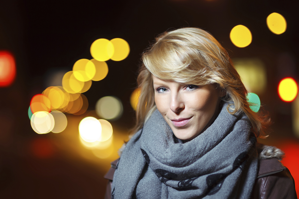 a portrait of a young woman wearing a scarf with blurred city lights behind her creating a bokeh effect 