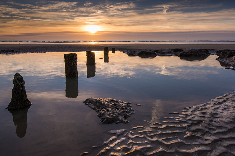 A landscape shot of rocks peering out of a very still and reflective rock pool as the sunsets in the distance painting the sky orange, lavender and pink as it sinks below the horizon 