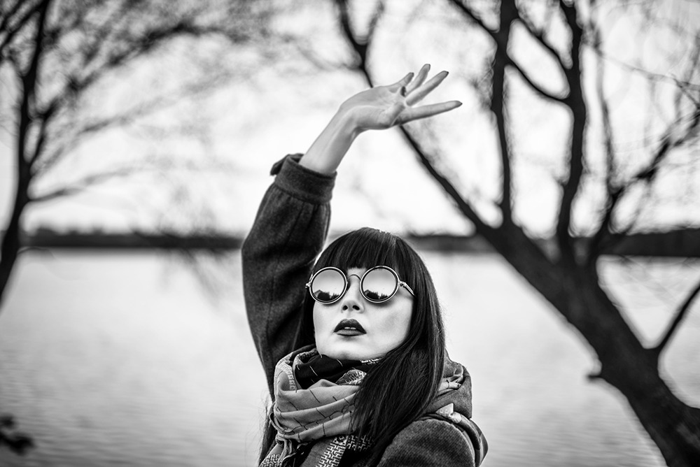 A portrait of a woman with arms raised as if she were dancing, a sharp cut fringe and circular sunglasses with deep black lenses