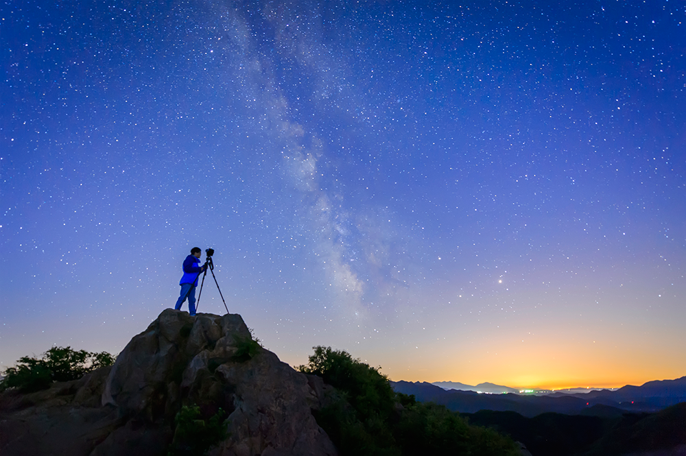 a man stands atop a small mountain with his tripod, the photo has been taken at night and the sky looks a deep purple with all of the stars shining through 