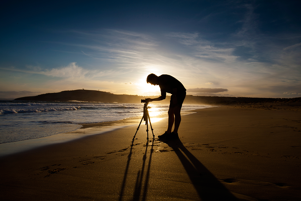  man takes a photo near the surf as the sun sets