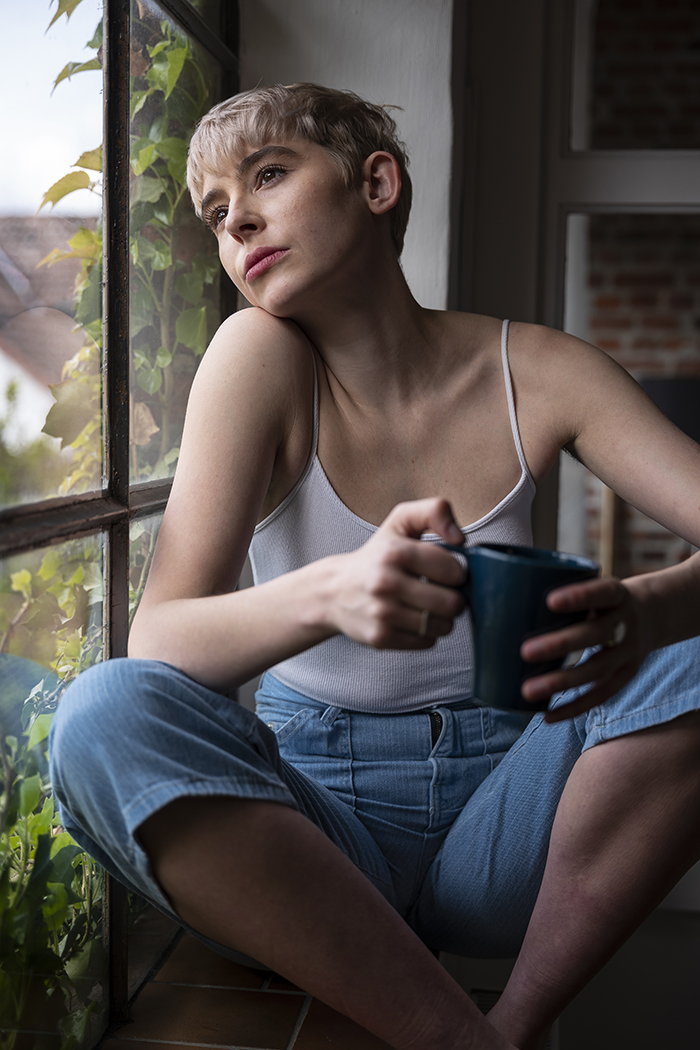 woman sat in window sill, looking out at window with a drink